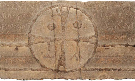 In the centre is the monogram of Christ, sculpted in a circle. The Greek cross with flared arms divides the circle into four segments. Christ is represented by the vertical arms of the cross forming a Greek c, with a loop on the upper arm forming a Greek P placed so as to be read from right to left, as in Arabic and Syriac. In the right and left upper segments, there appear the sun and the moon – a broken sun and a moon full like the breast of a woman, figuring the cosmic signs that accompanied the death of Jesus: full moon (the date of Pasch), and the darkening of the sun (Lk 23.44). In the bottom segments, one recognises the first and last letters of the Greek alphabet: Alpha and Omega, placed in the direction of reading in Syriac and Arabic, from right to left, suspended by curious straps to the arms of the cross. They represent Christ, beginning and end of all things (Ap 1.8), suspended on the gibbet.